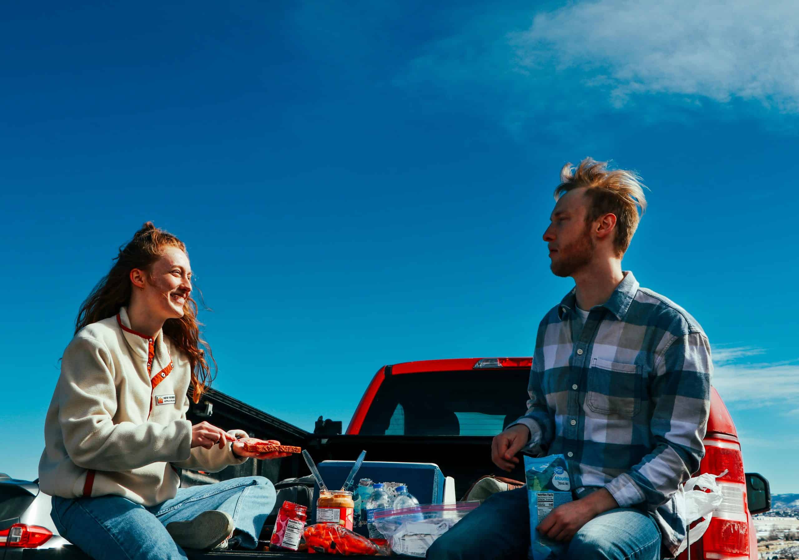 A cheerful couple having a picnic on a truck tailgate under clear blue skies. game day