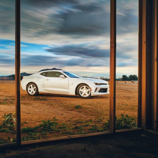 A white Camaro parked in an open field, viewed from a rustic window frame at sunset. chevy camaro