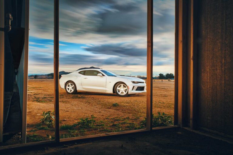A white Camaro parked in an open field, viewed from a rustic window frame at sunset. chevy camaro