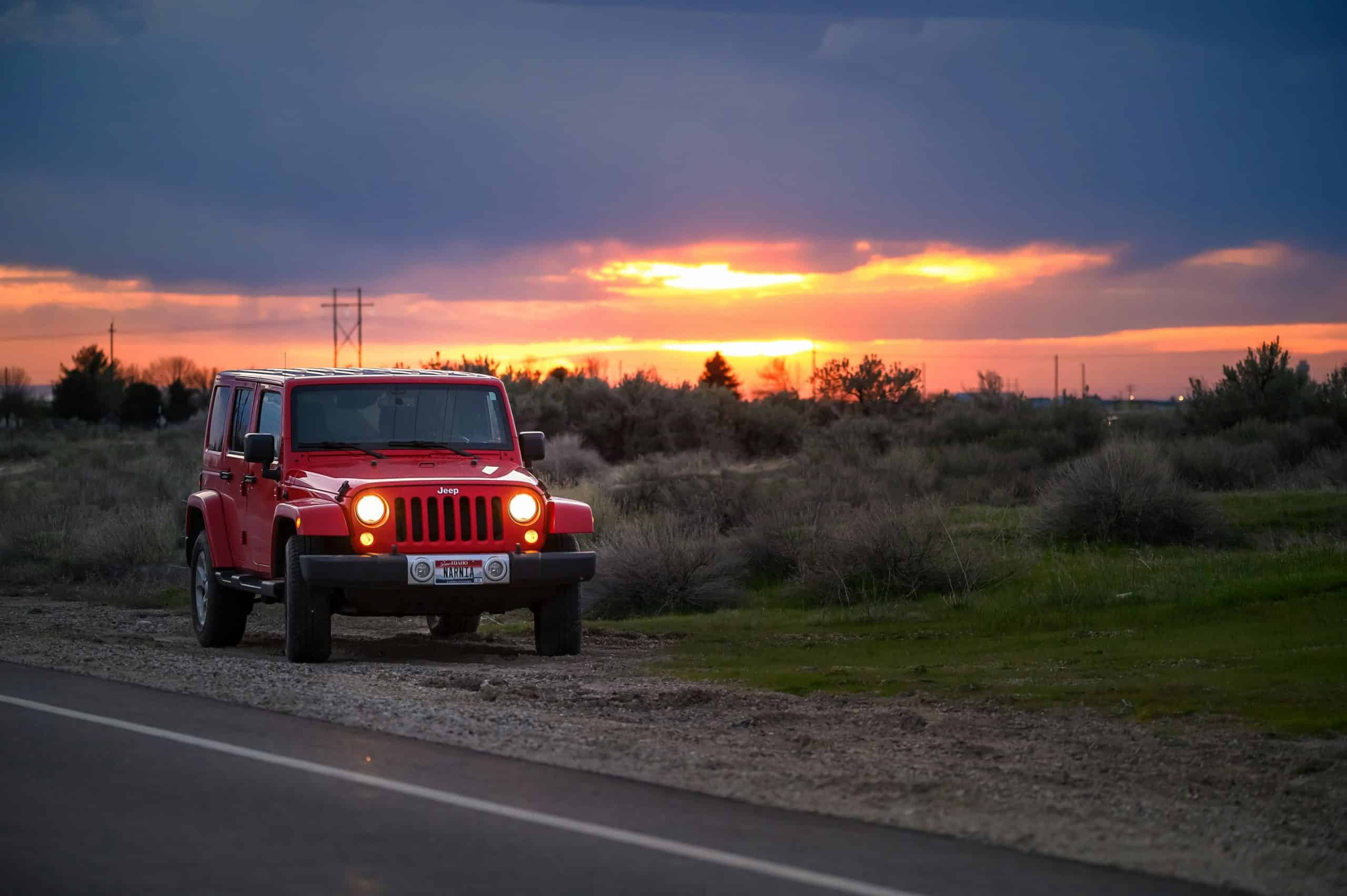 A red Jeep parked beside a rural road at sunset, with a striking sky in the background.