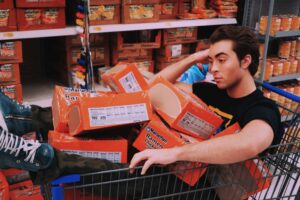 A man lounging in a shopping cart filled with Maruchan ramen in a supermarket aisle. ultra-processed foods