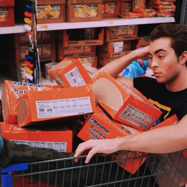 A man lounging in a shopping cart filled with Maruchan ramen in a supermarket aisle. ultra-processed foods