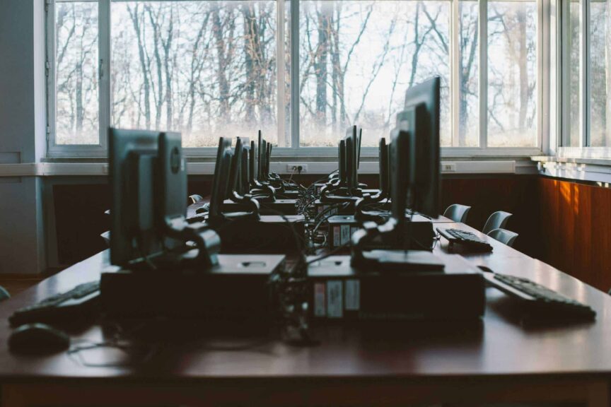 An empty computer lab with multiple workstations and large windows during the daytime. pc/intel