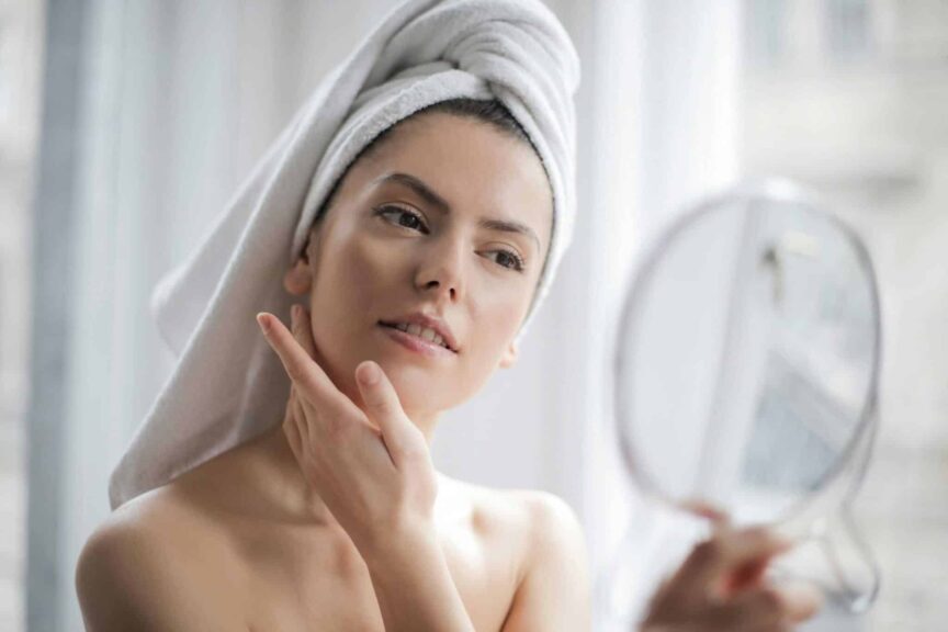 This photograph features a woman at home looking in a mirror. A home facial steamer can be part of a skincare routine.
