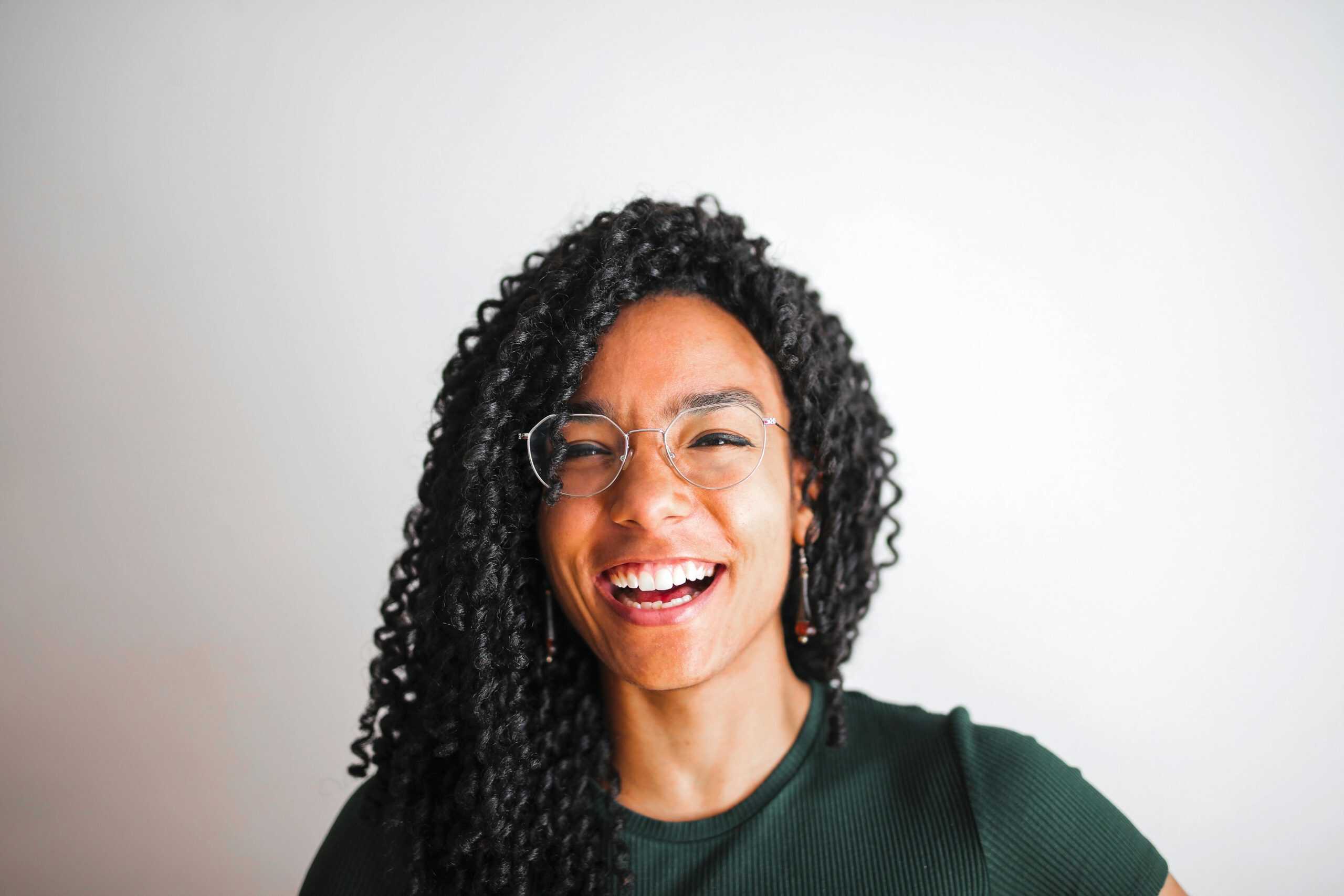 A joyful portrait of a young woman with glasses and curly hair smiling against a simple white background, daily devotion