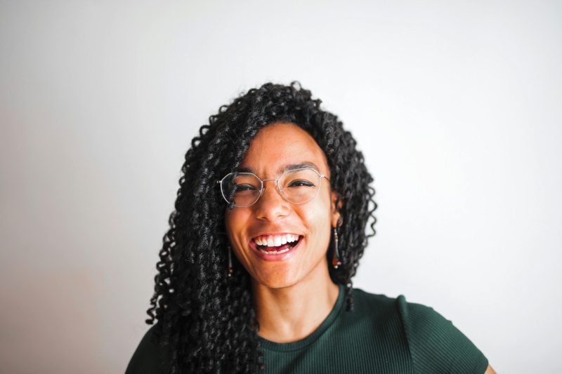 A joyful portrait of a young woman with glasses and curly hair smiling against a simple white background. daily devotion