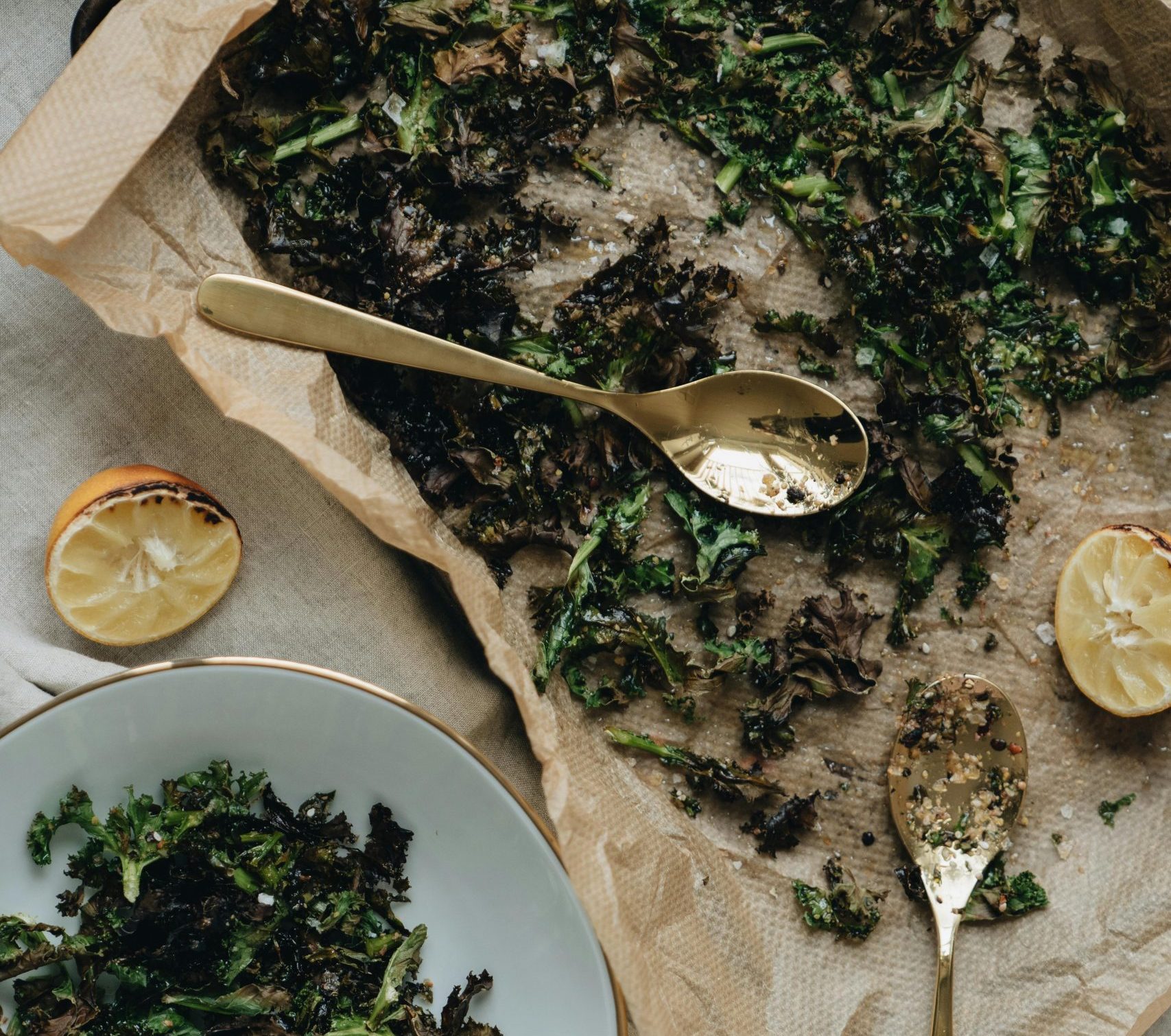 Top view of baked kale chips on baking paper with lemons and spoons.