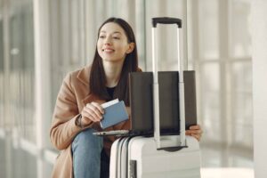 A woman sitting with her laptop and luggage, holding a passport, waiting in an airport terminal. holiday flights