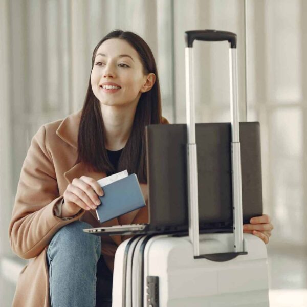 A woman sitting with her laptop and luggage, holding a passport, waiting in an airport terminal. holiday flights