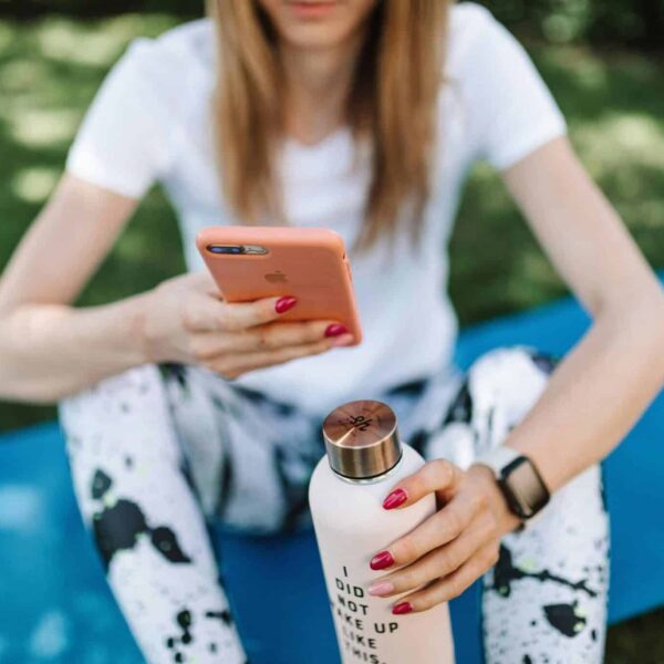spirituality, technology, Woman using smartphone while holding bottle on yoga mat outdoors.