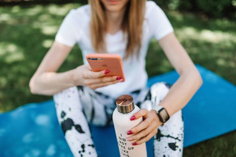 spirituality, technology, Woman using smartphone while holding bottle on yoga mat outdoors.