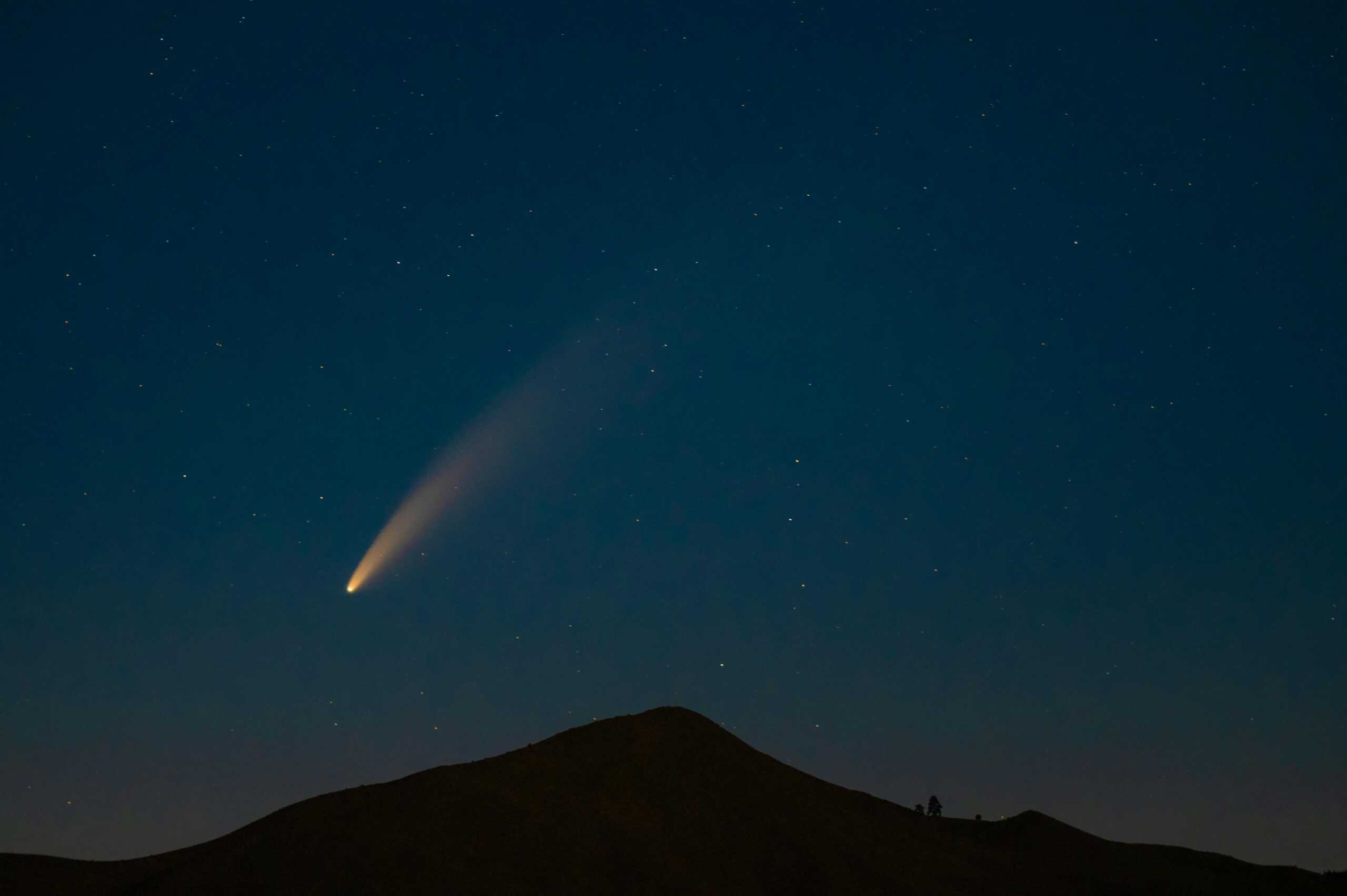 A breathtaking capture of a comet streaking across a star-filled night sky above a mountain silhouette.