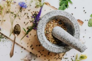 Top view of herbs and spices prepared for cooking aromatics, being ground with a stone mortar and pestle on a wooden cutting board.