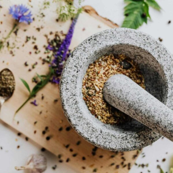 Top view of herbs and spices prepared for cooking aromatics, being ground with a stone mortar and pestle on a wooden cutting board.