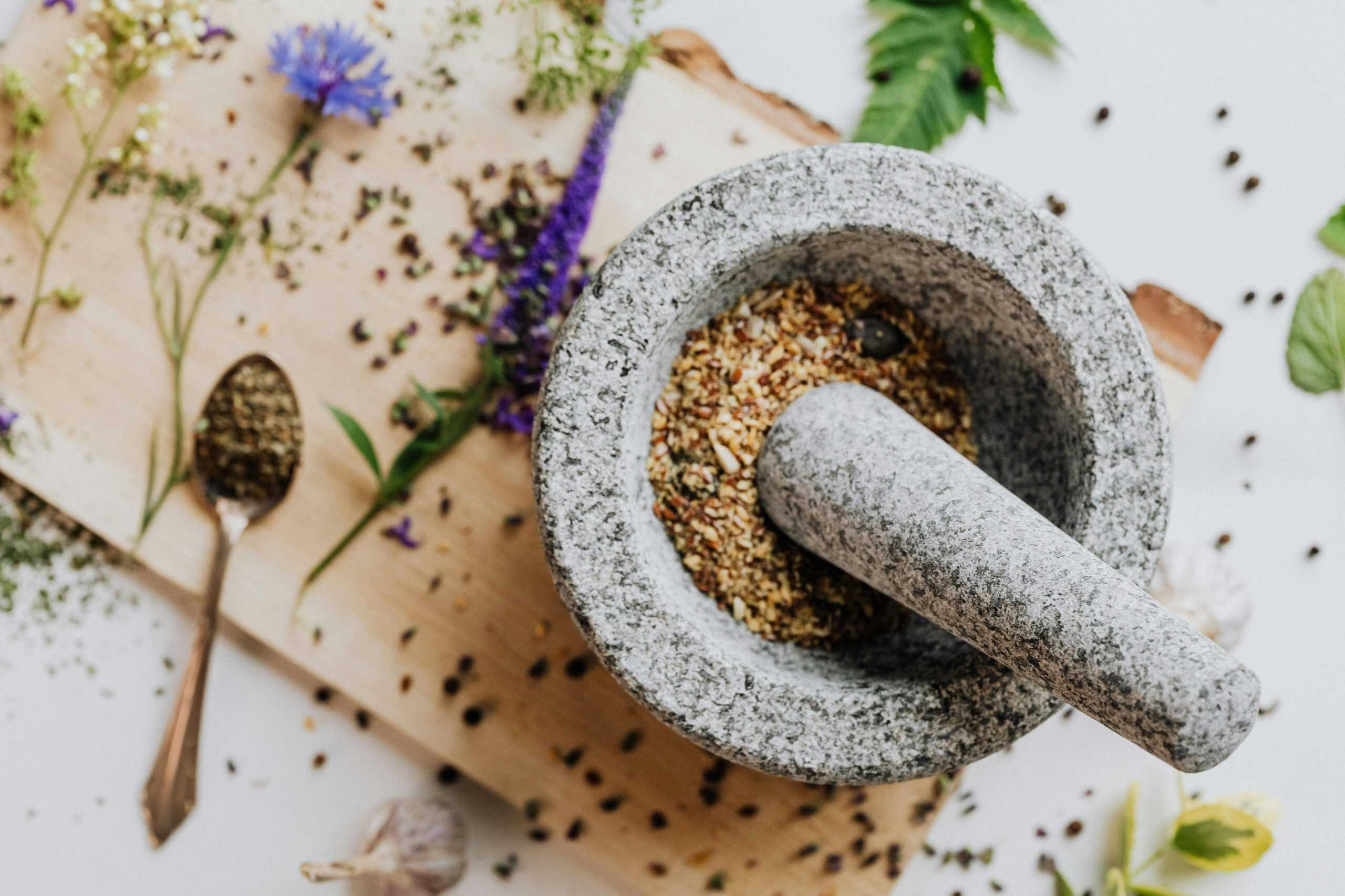 Top view of herbs and spices prepared for cooking aromatics, being ground with a stone mortar and pestle on a wooden cutting board.