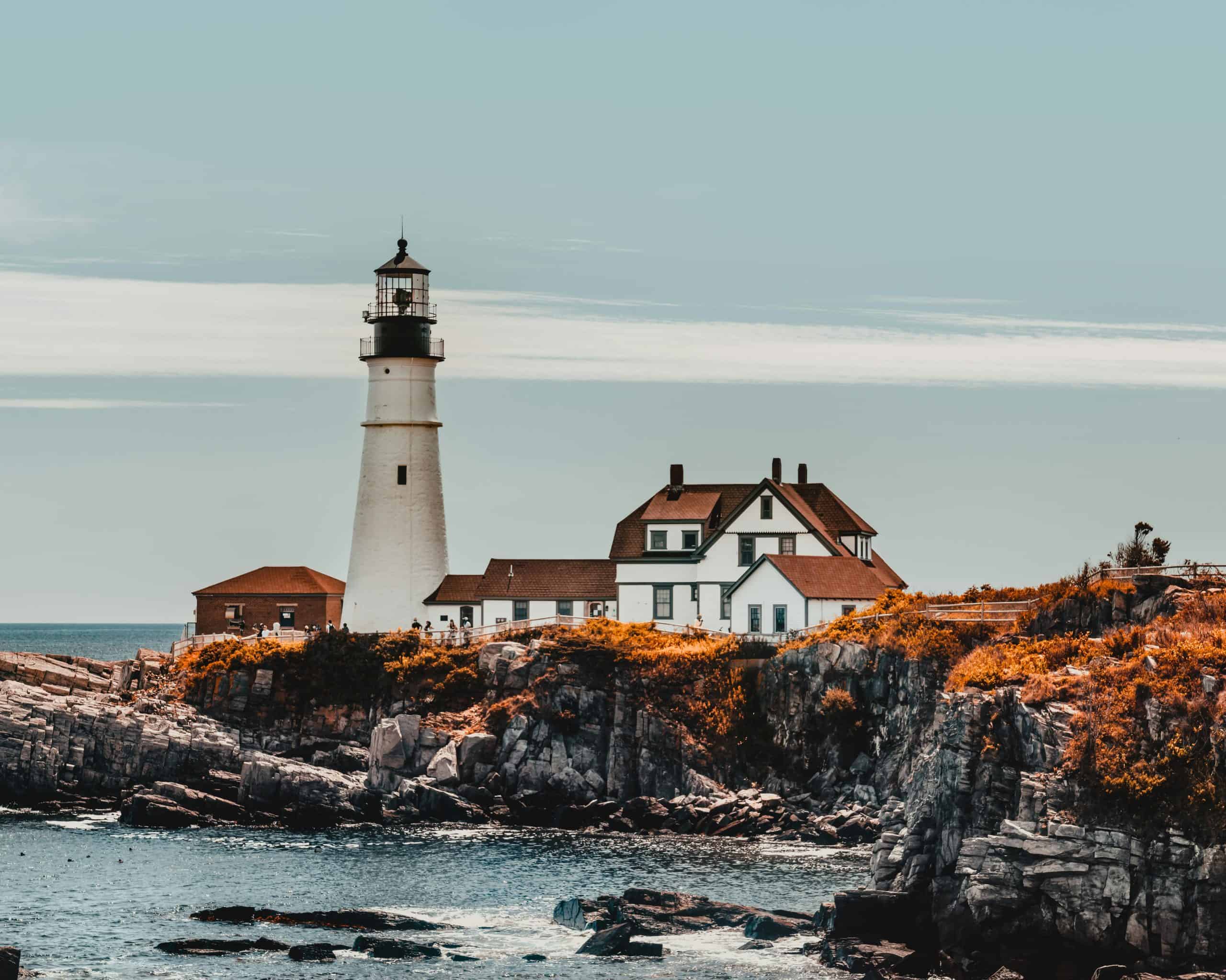 Lighthouse near big house near orange grass on rocky shore near ocean under bright sky in daytime