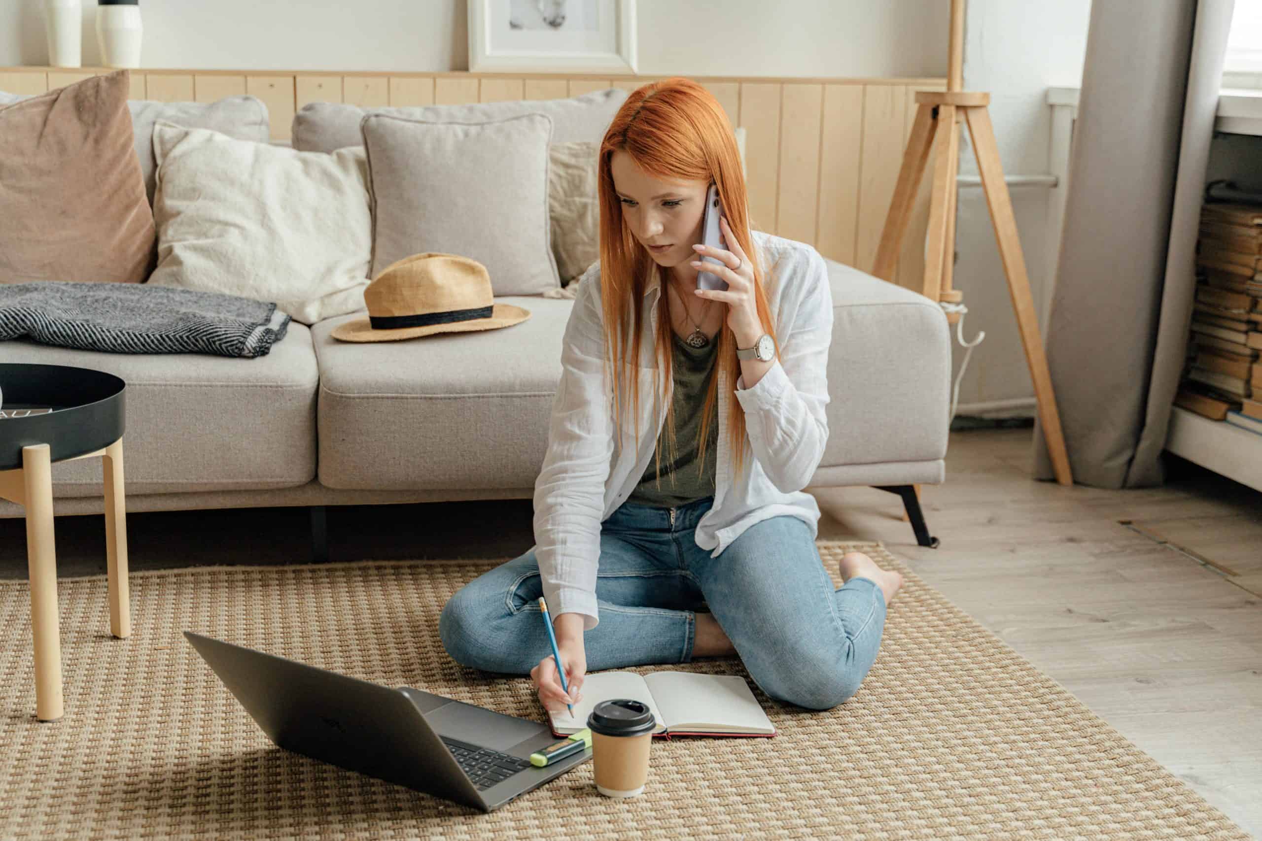A woman sitting on the floor using a laptop and phone in a modern home office environment.