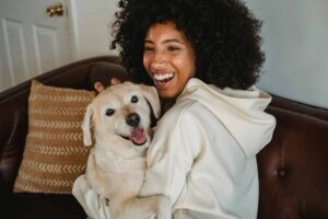 Happy young black lady hugging with dog while sitting on sofa in casual outfit in room. pets in dorms