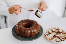 Close-up of hands adding chocolate glaze to a Bundt cake beside festive cookies.
