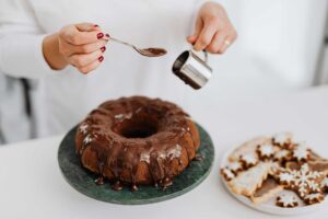 Close-up of hands adding chocolate glaze to a Bundt cake beside festive cookies.