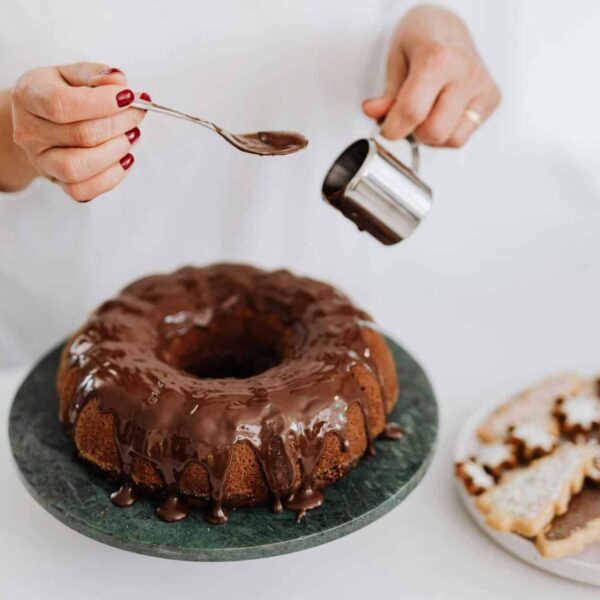 Close-up of hands adding chocolate glaze to a Bundt cake beside festive cookies.