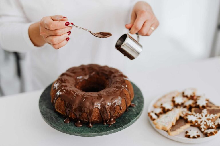 Close-up of hands adding chocolate glaze to a Bundt cake beside festive cookies.