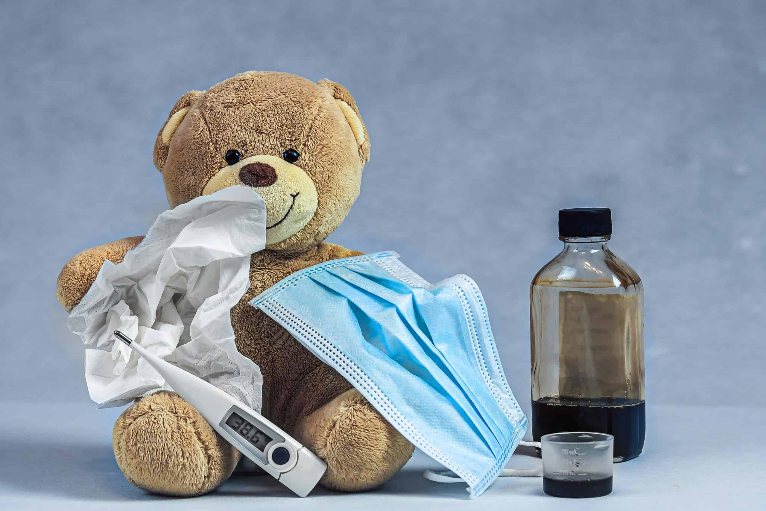 A teddy bear with cough syrup, a mask, thermometer, and tissue highlights an illness concept.