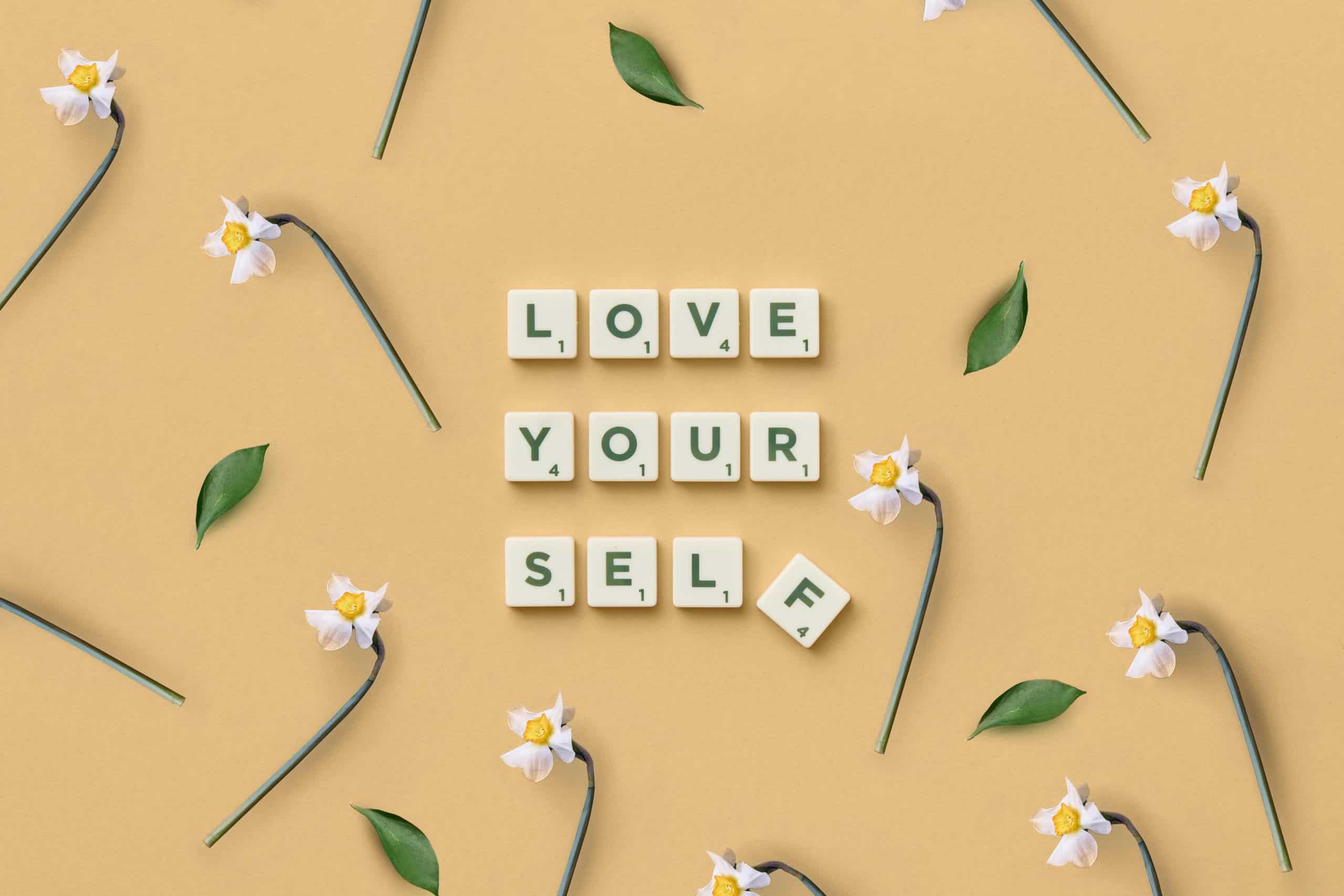 Scrabble tiles spelling 'Love Yourself' surrounded by flowers and leaves on a flat surface.