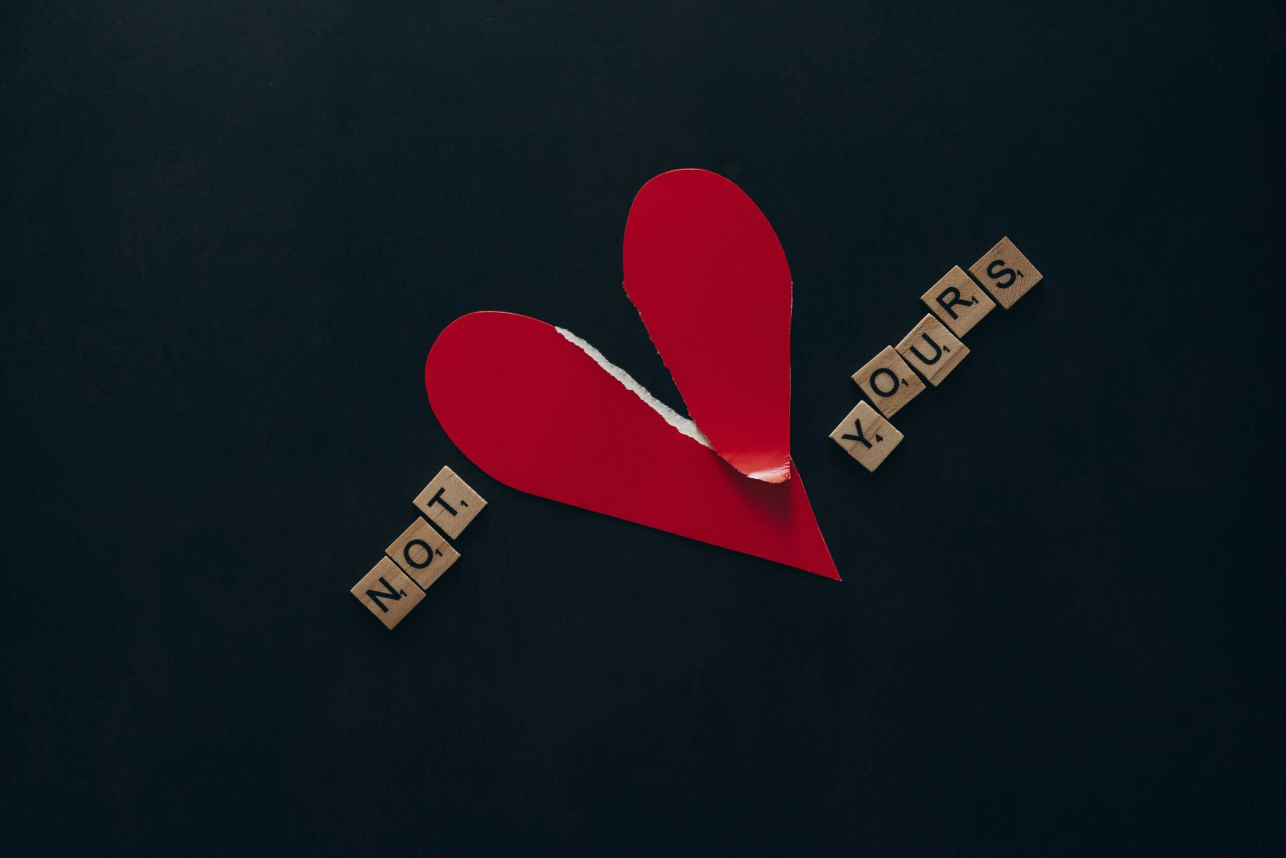 Conceptual image of a torn red heart with Scrabble tiles spelling 'NOT YOURS' on a dark background.
