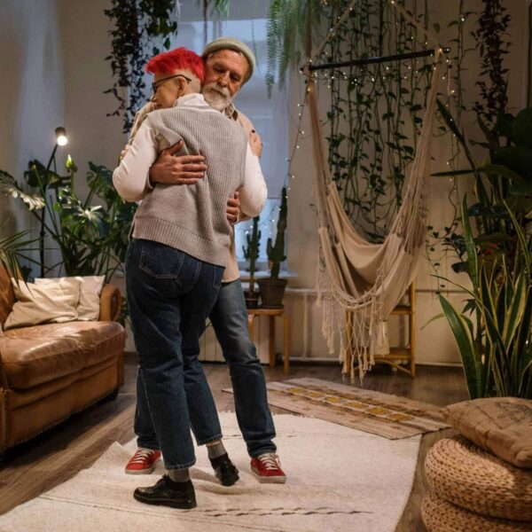 An elderly couple shares a tender embrace in a tranquil, plant-filled living room setting. accessibility