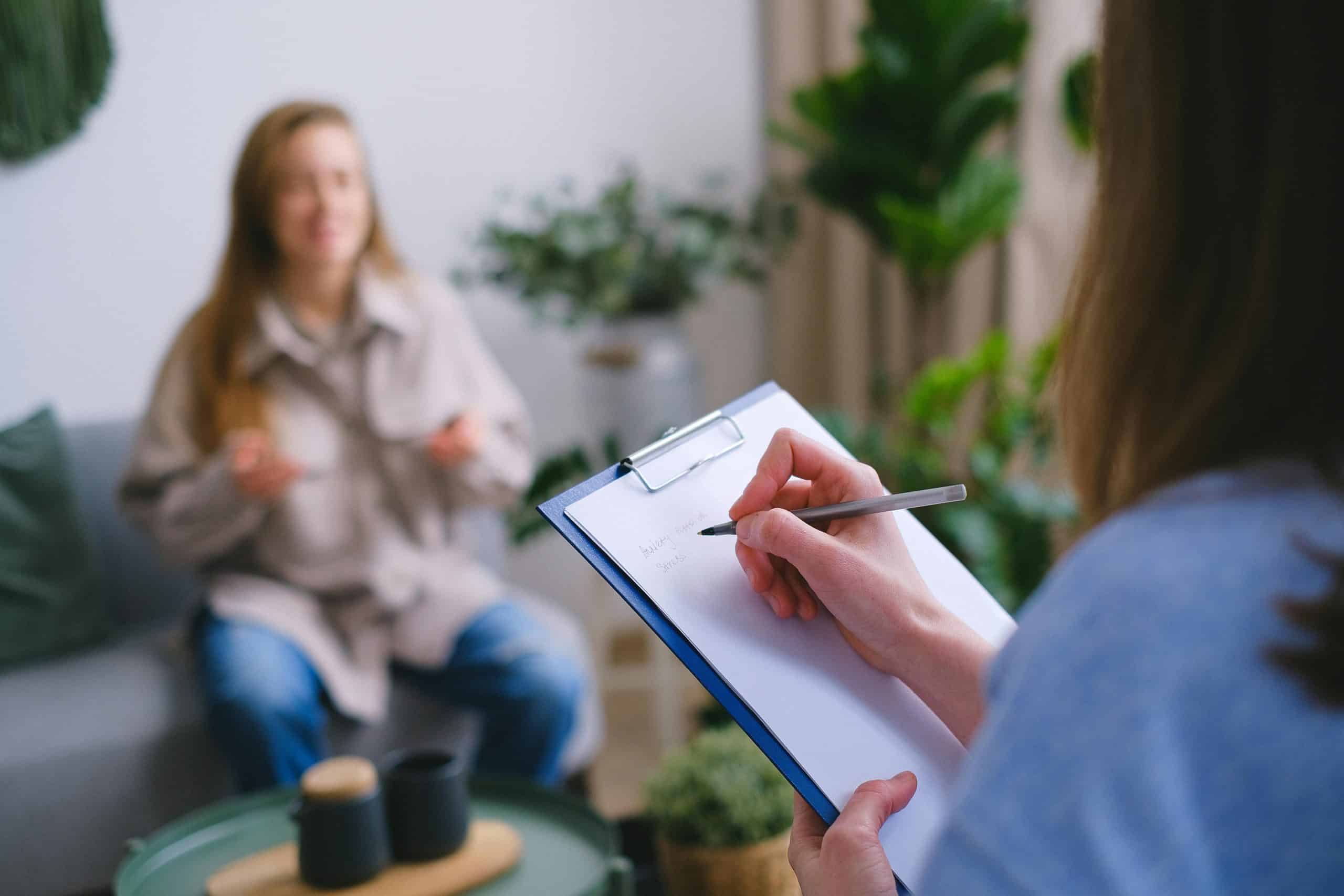 Unrecognizable professional female psychologist writing on clipboard while sitting against client on blurred background during psychotherapy session in light office, inner child, relationships