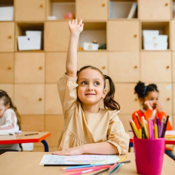 Three children engaged in learning activities inside a classroom, with one raising her hand, back-to-school, immunizations
