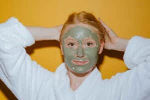 Woman enjoying self-care with a green face mask against a yellow background.