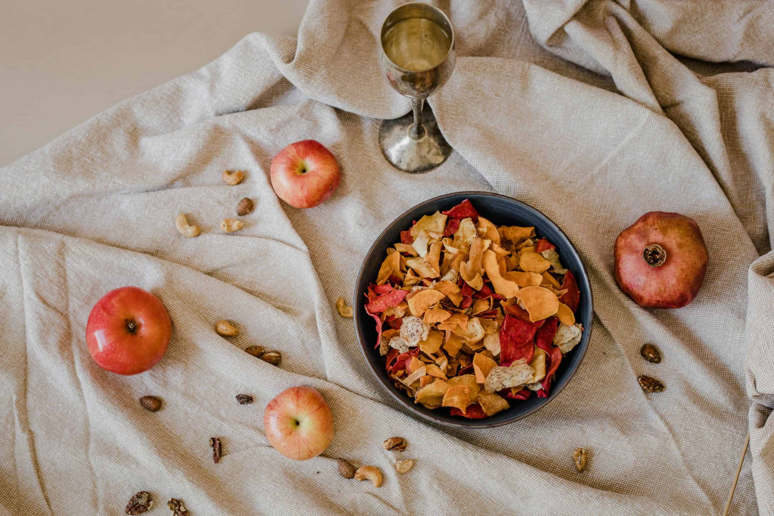 Overhead view of a bowl of dried fruit with apples, nuts, and a silver goblet on a rustic fabric.