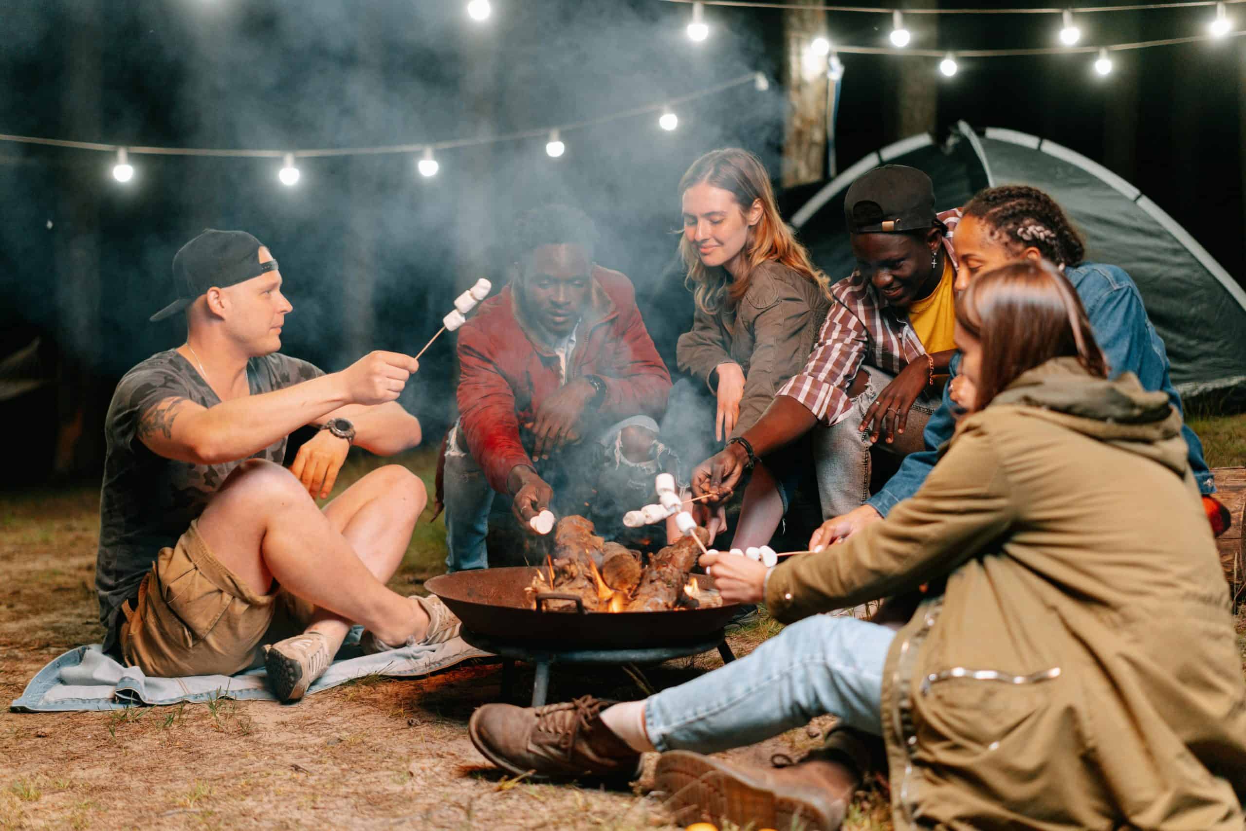 A group of friends relaxes by a bonfire, roasting marshmallows under string lights on a camping trip.