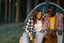 Two friends enjoying a cheerful outdoor camping trip, sitting on a log, smiling with beer bottles. Fall camping, cool-weather