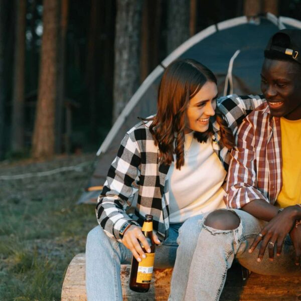Two friends enjoying a cheerful outdoor camping trip, sitting on a log, smiling with beer bottles. Fall camping, cool-weather