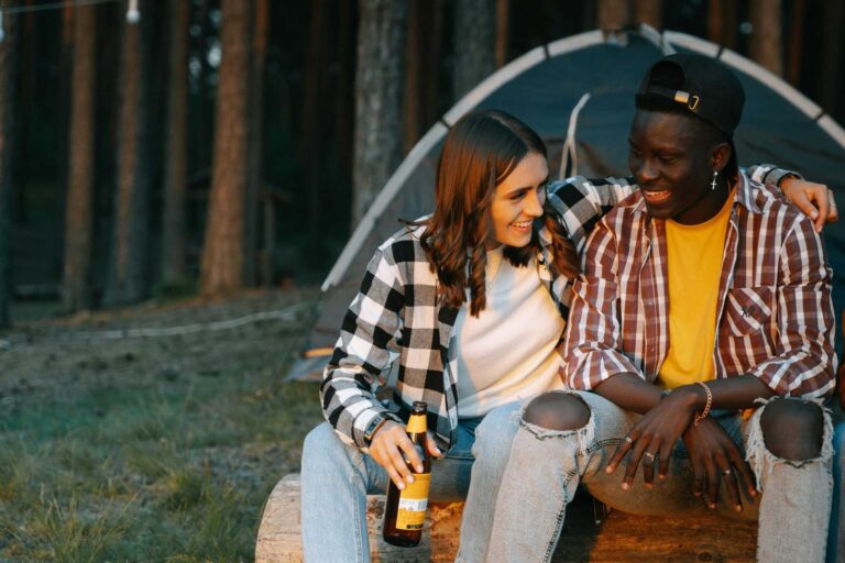 Two friends enjoying a cheerful outdoor camping trip, sitting on a log, smiling with beer bottles. Fall camping, cool-weather