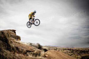 Challenge travel, adventure travel, A cyclist performs a daring jump off a rocky cliff under a cloudy sky.
