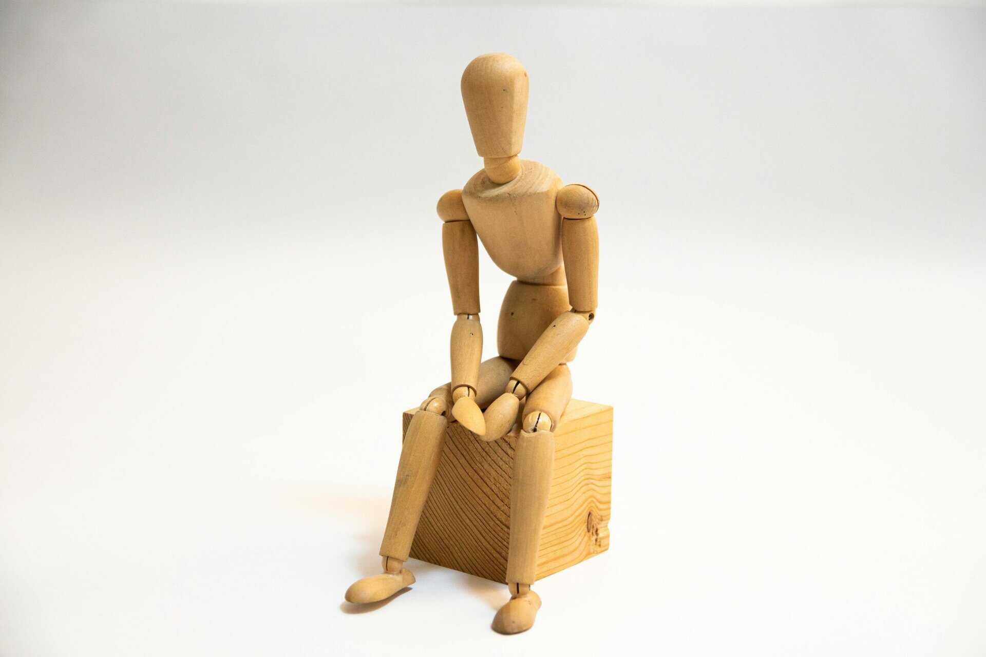 A wooden artist's mannequin seated on a wooden block against a neutral background.