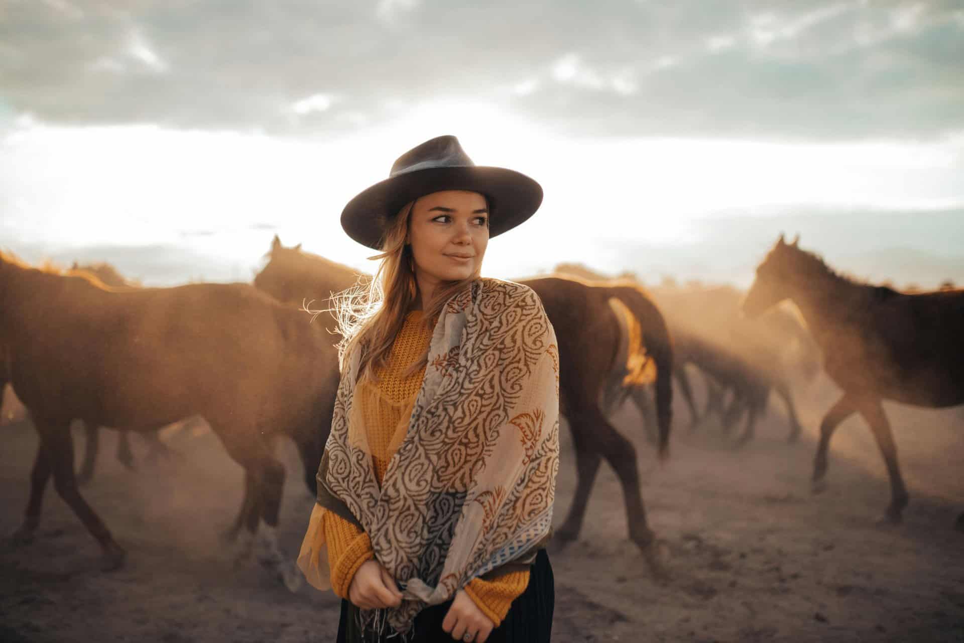 Woman in a fedora stands amidst a herd of horses at sunset, exuding a serene and adventurous vibe. Fall fashion, fashion trend