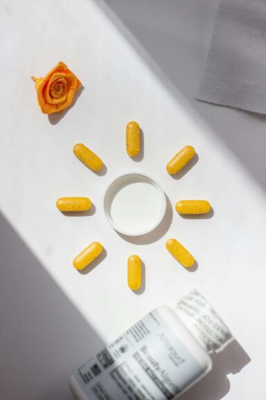 Bright yellow vitamin pills arranged in a sun shape beside a bottle and rose accent, visually representing ingredients often found in skin serum formulations.
