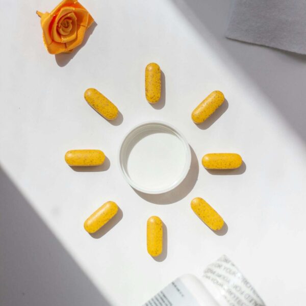 Bright yellow vitamin pills arranged in a sun shape beside a bottle and rose accent, visually representing ingredients often found in skin serum formulations.