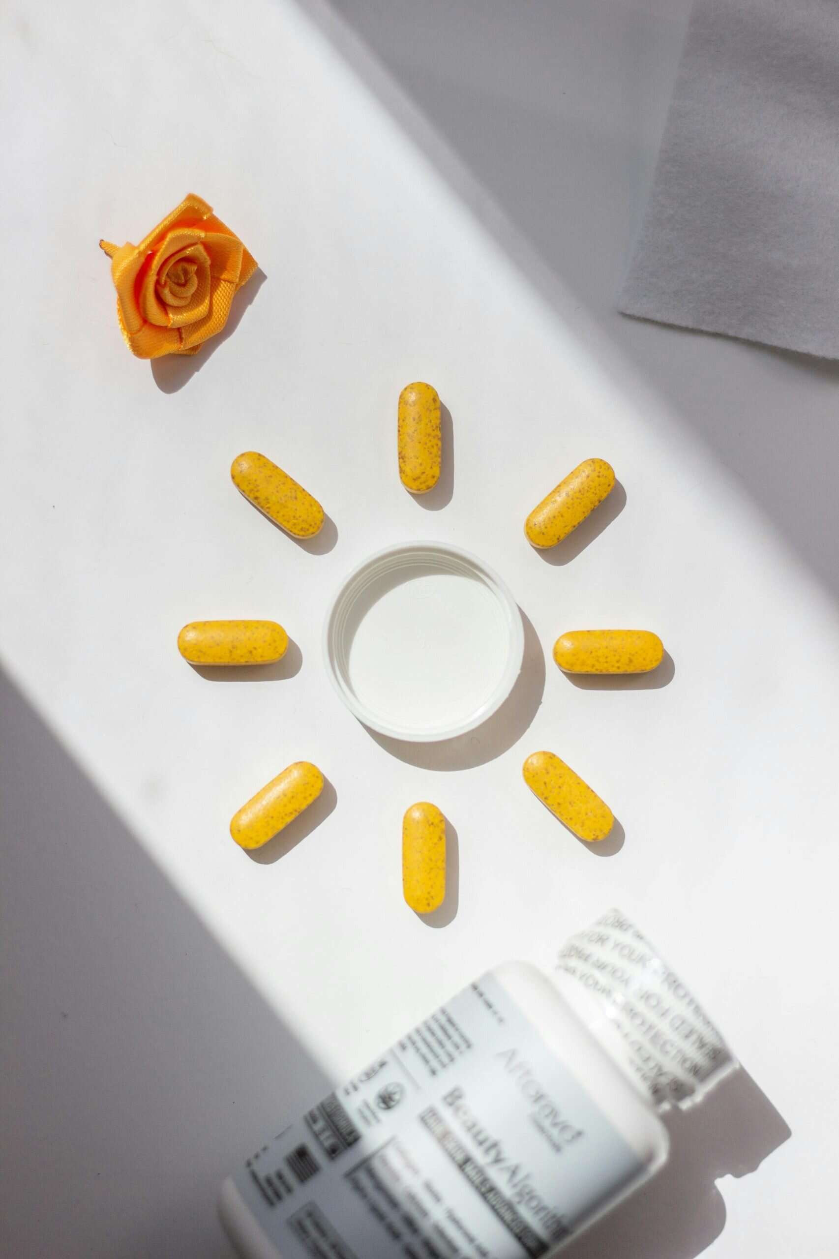 Bright yellow vitamin pills arranged in a sun shape beside a bottle and rose accent, visually representing ingredients often found in skin serum formulations.