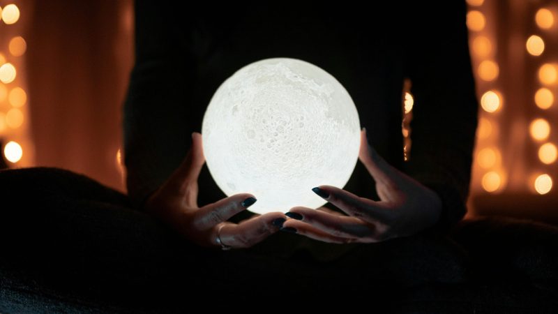 A person holding a glowing moon lamp, surrounded by warm bokeh lights, creating a mystical indoor atmosphere.