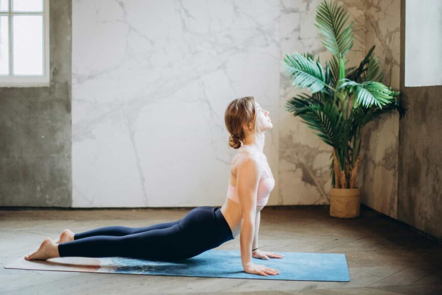 Woman in yoga pose on mat, exercising indoors with natural light and plant decor, somatic psychology, healing