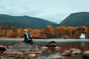 Ireland, fall travel, A woman seated on a rock, enjoying the tranquil autumn landscape by a lake with mountains in Ireland.