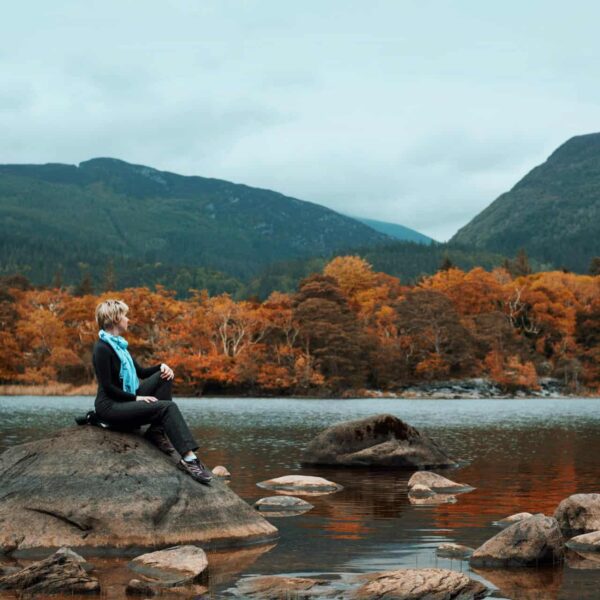 Ireland, fall travel, A woman seated on a rock, enjoying the tranquil autumn landscape by a lake with mountains in Ireland.
