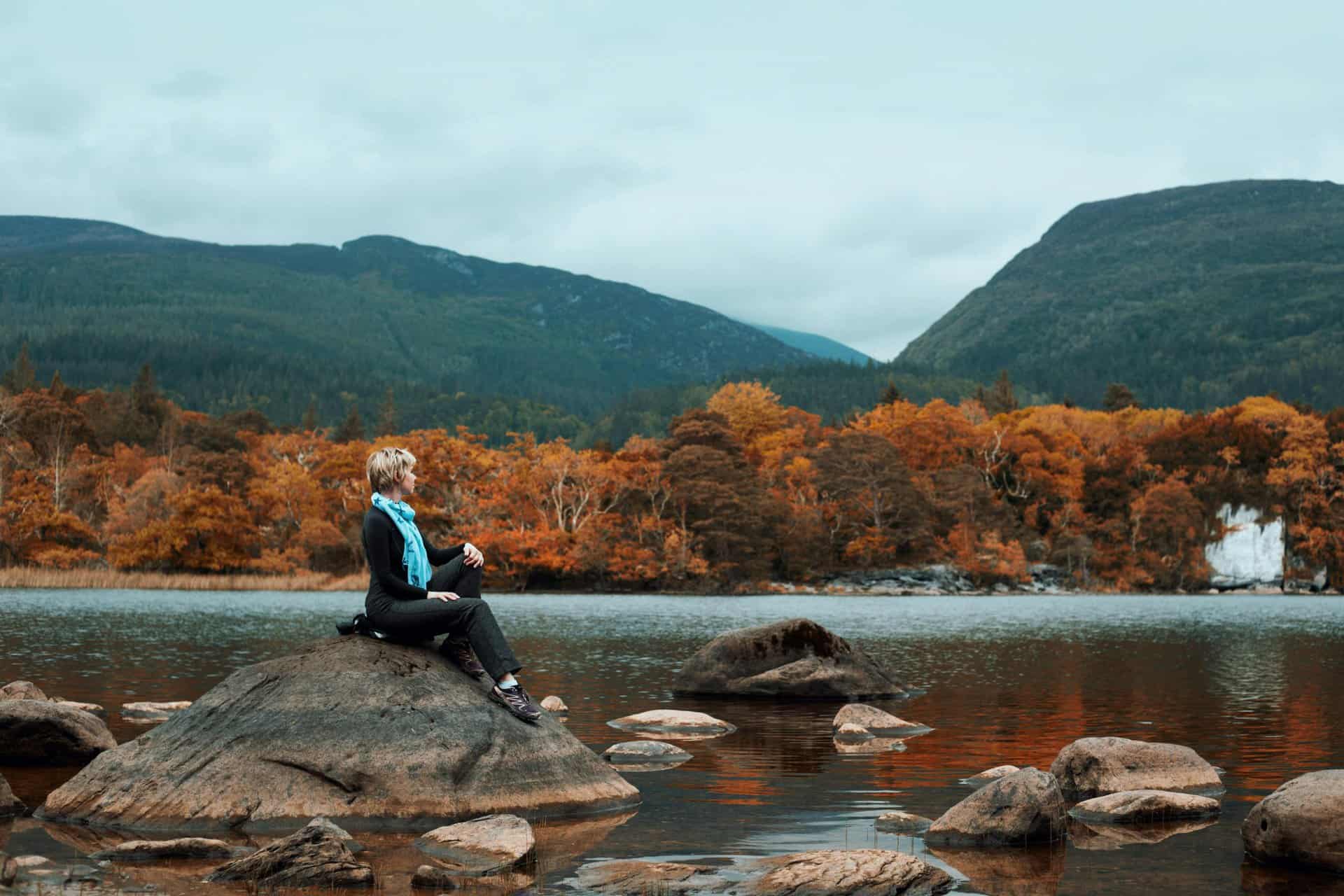 Ireland, fall travel, A woman seated on a rock, enjoying the tranquil autumn landscape by a lake with mountains in Ireland.
