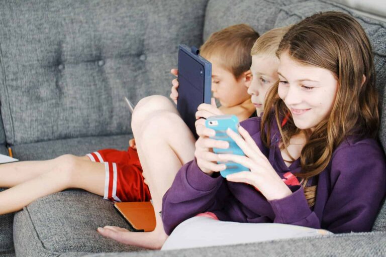 Three children sitting on a couch, engaged with electronic devices, smiling and enjoying leisure time, devices and screen time.